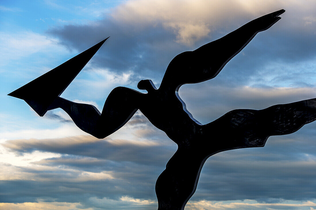 Silhouettierte Skulptur einer Person, die im Begriff ist, ein Papierflugzeug namens Utkast zu werfen, von Kare Groven, auf dem Flughafen Gardermoen vor einem Gemisch aus grauen Wolken und blauem Himmel kurz nach Sonnenuntergang; Akershus, Norwegen