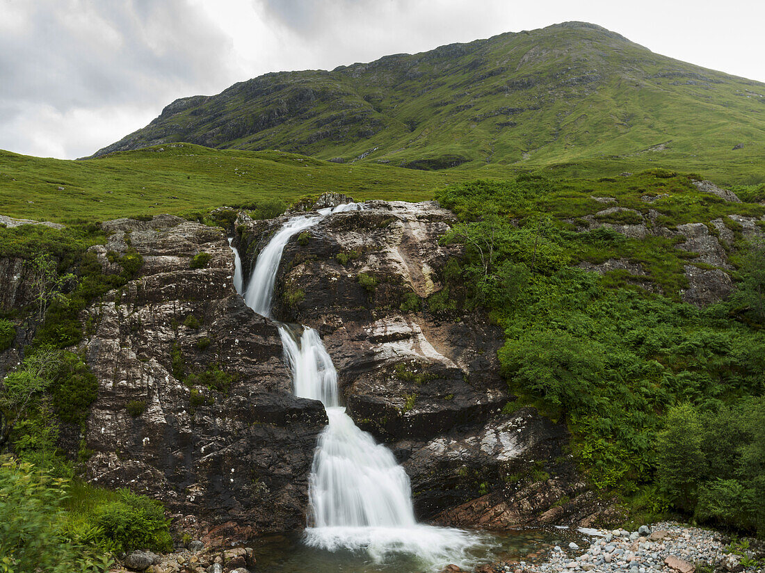 Wasserfall über einer zerklüfteten Felsklippe mit üppigem, grünem Gras in der Landschaft unter einem bewölkten Himmel; Schottland