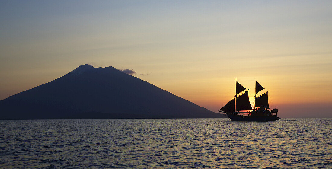 Blick auf den Ozean mit einer Yacht mit Segeln und einem Vulkan, der sich gegen die untergehende Sonne abhebt