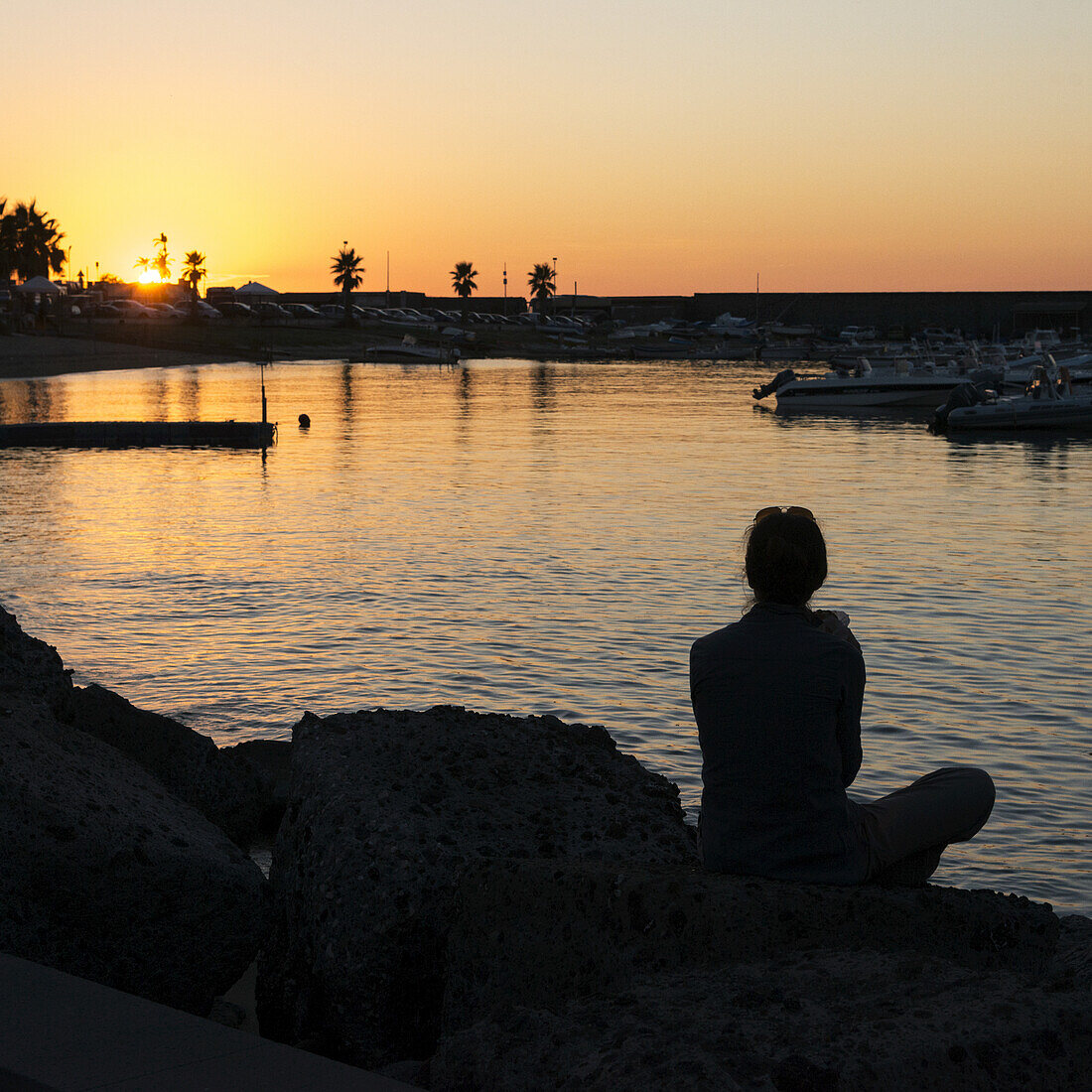 Silhouette einer Frau, die am Ufer sitzt und bei Sonnenuntergang über einen Hafen blickt; Forio, Ischia, Kampanien, Italien