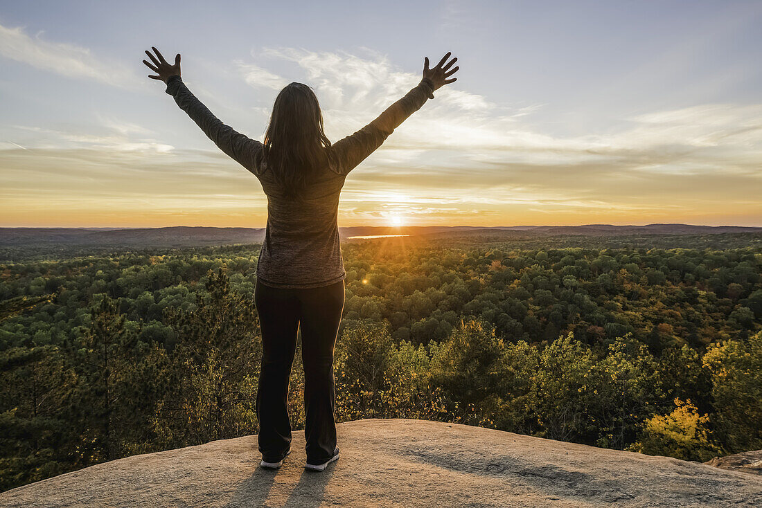 Frau auf einer Klippe stehend mit ausgebreiteten Händen bei Sonnenuntergang, Algonquin Park; Ontario, Kanada