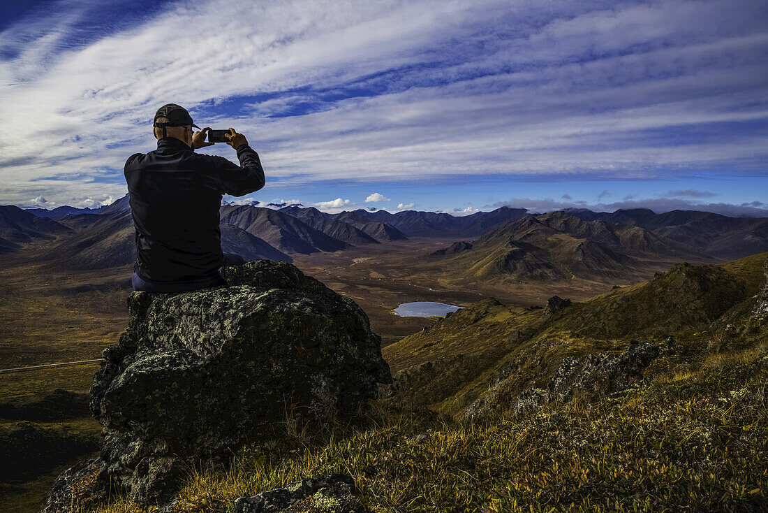 Mann, der mit einem Smart Phone Fotos auf einem Aussichtspunkt mit Blick auf das Blackstone Valley entlang des Dempster Highway macht; Yukon, Kanada