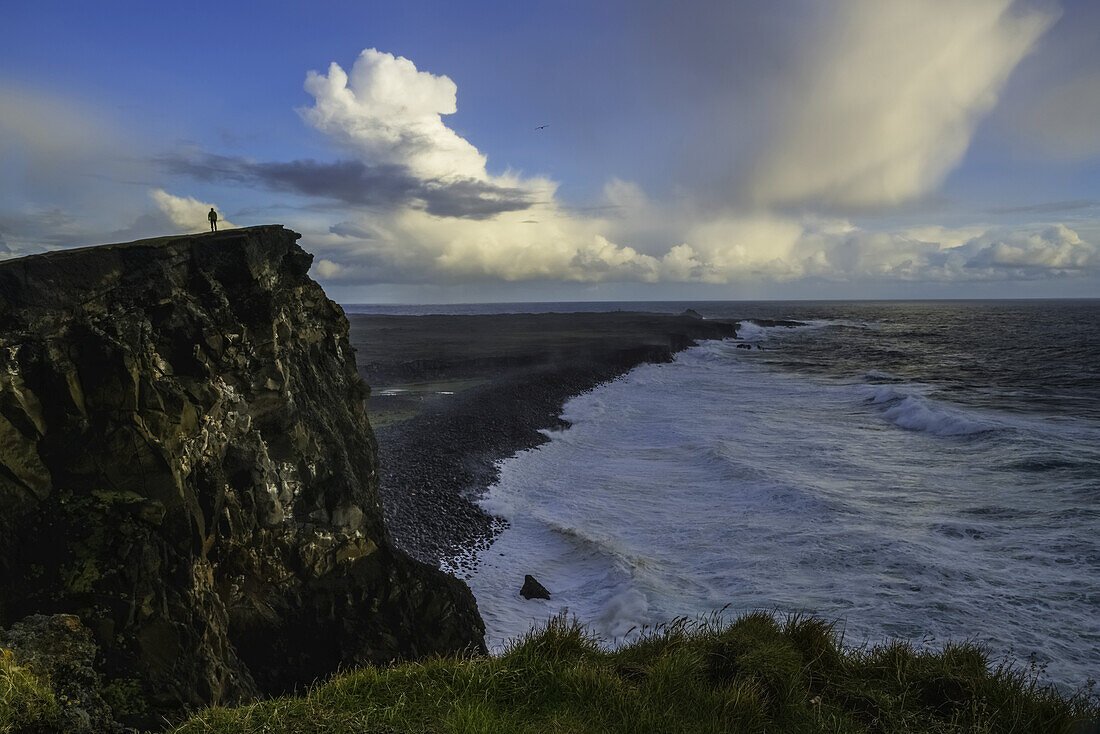 Person Standing On A Cliff Face While The Atlantic Ocean Pounds The Shores Beneath Them; Iceland