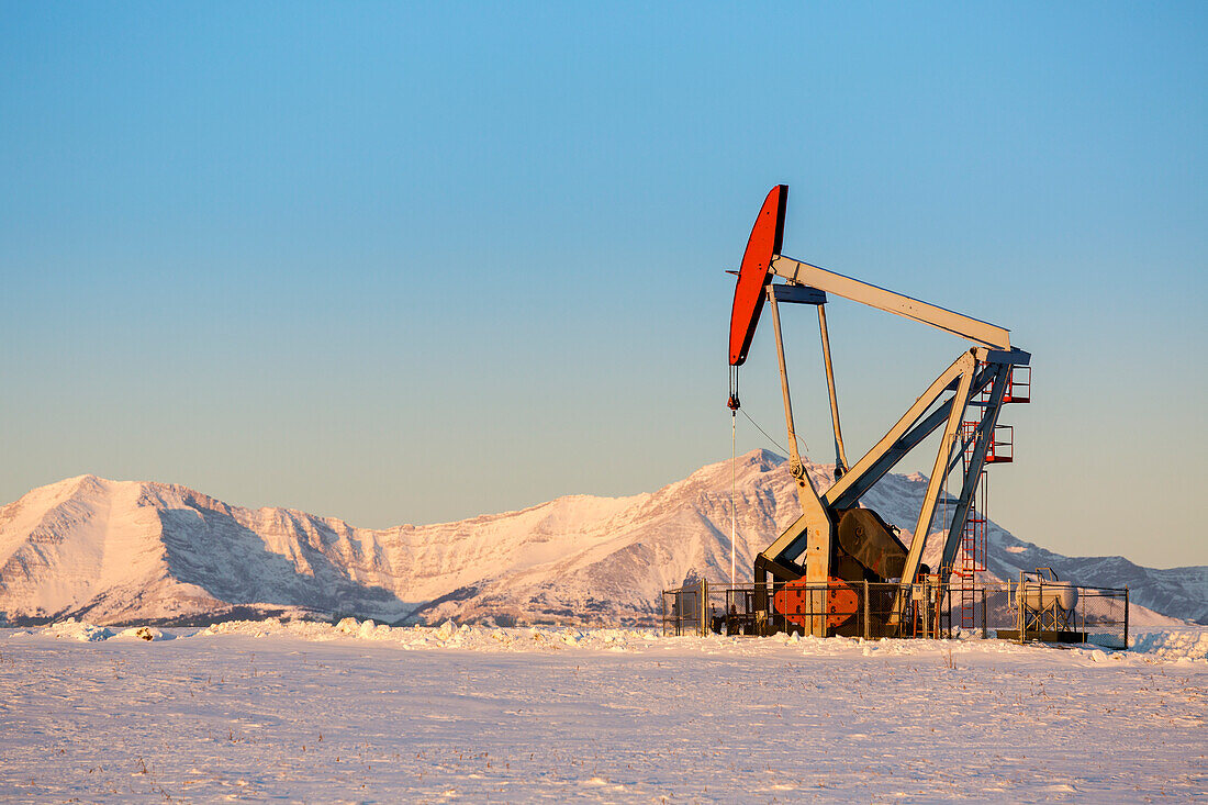 Pumpjack with the warm light at sunrise in a snow-covered field with snow-covered mountains and blue sky in the background; Longview, Alberta, Canada