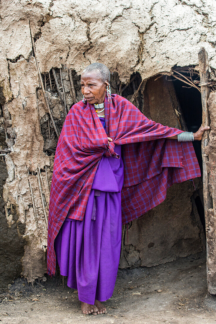 Ältere Maasai-Frau in traditioneller Kleidung steht am Eingang ihrer Lehmhütte im Ngorongoro-Schutzgebiet; Tansania