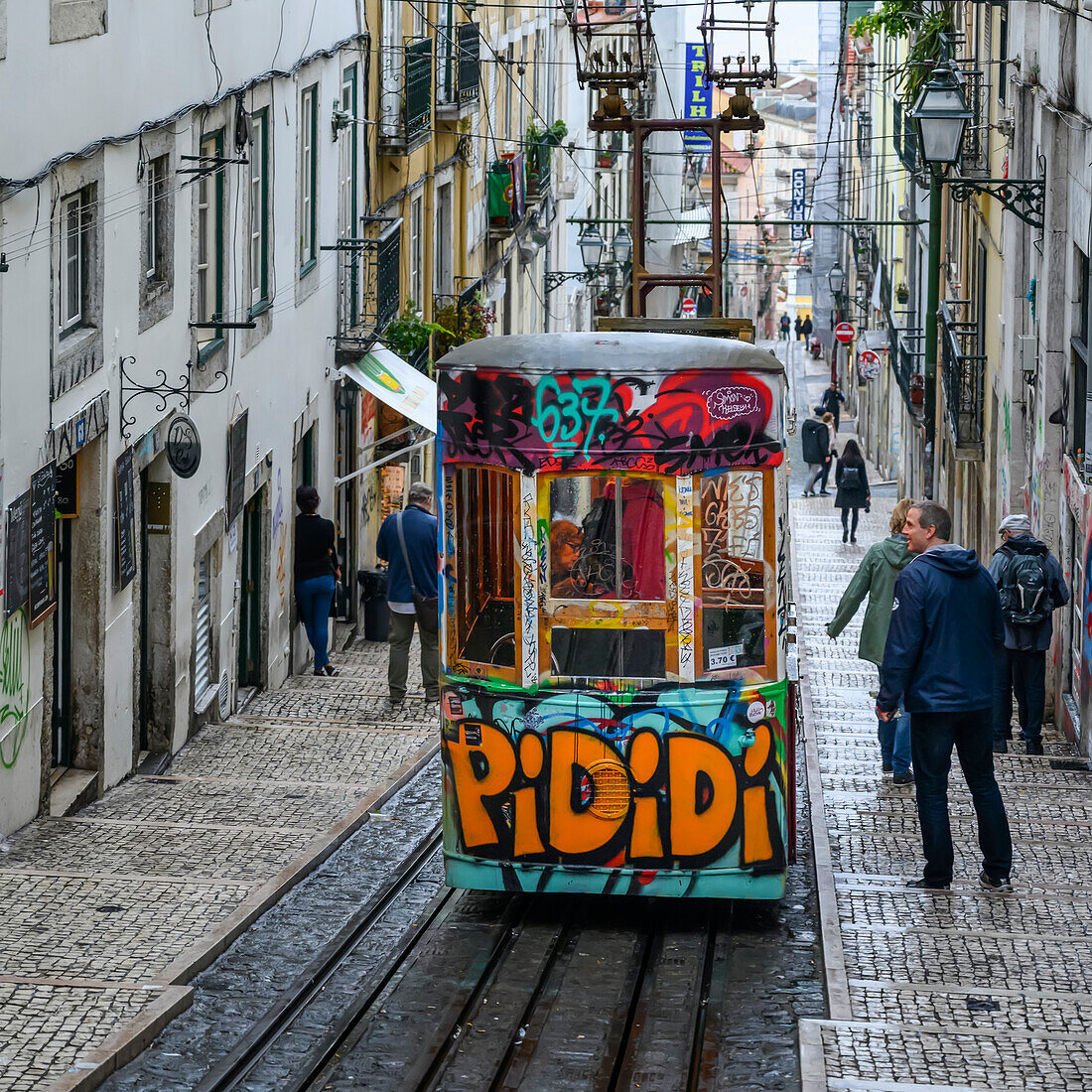 Straßenbahn mit Graffiti, die auf einer abschüssigen Straße mit Fußgängern und Wohngebäuden bergab fährt; Lissabon, Region Lisboa, Portugal