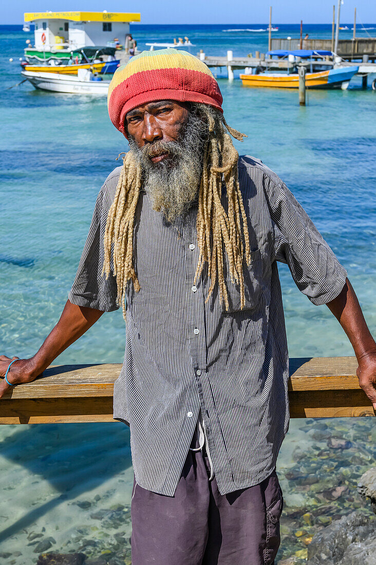 A man stands along the water's edge posing for the camera with boats moored at the dock in the background, depicting the people and lifestyle on the island of Roatan, West End Village; Roatan, Bay Islands Department, Honduras