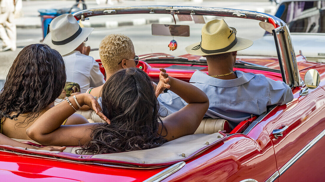 Three woman and two men ride in a red convertible car; Havana, Cuba