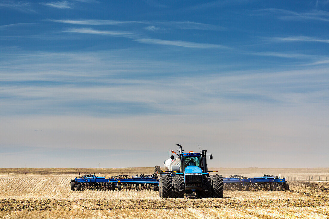 Tractor with air seeder, seeding a stubble field with blue sky and hazy clouds, near Beiseker; Alberta, Canada