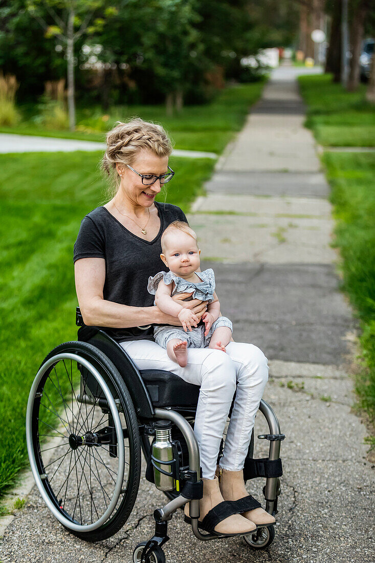 Eine querschnittsgelähmte Mutter, die ihr Baby auf dem Schoß trägt, während sie einen Rollstuhl benutzt, an einem warmen Sommernachmittag im Freien: Edmonton, Alberta, Kanada