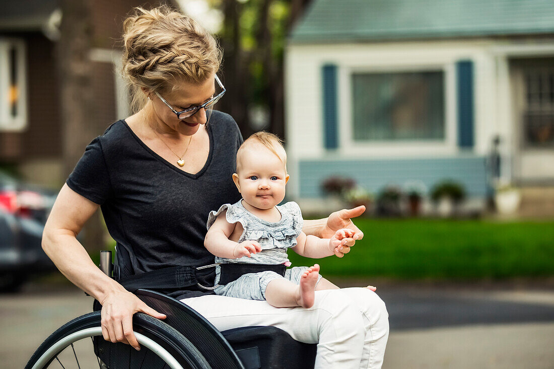 Eine querschnittsgelähmte Mutter, die ihr Baby auf dem Schoß trägt, während sie an einem warmen Sommernachmittag im Freien einen Rollstuhl benutzt: Edmonton, Alberta, Kanada
