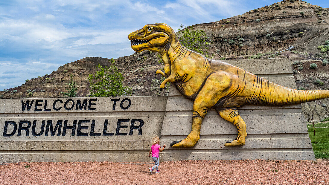 Großes Schild 'Welcome to Drumheller' mit einem Dinosaurier und einem jungen Mädchen, das darauf zuläuft; Drumheller, Alberta, Kanada