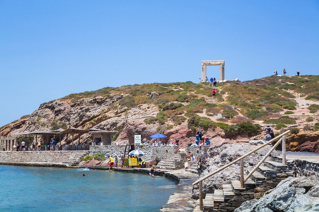 Tourists and swimmers enjoy a recreation area on the waterfront of the Mediterranean Sea with Temple of Apollo (Portara) in the background; Chora, Naxos Island, Cyclades, Greece; Greece