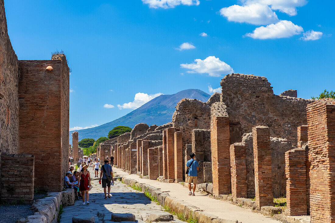 Tourists walk through the excavated ruins down a street in Pompeii with Mount Vesuvius in the background; Pompeii, Province of Naples, Campania, Italy