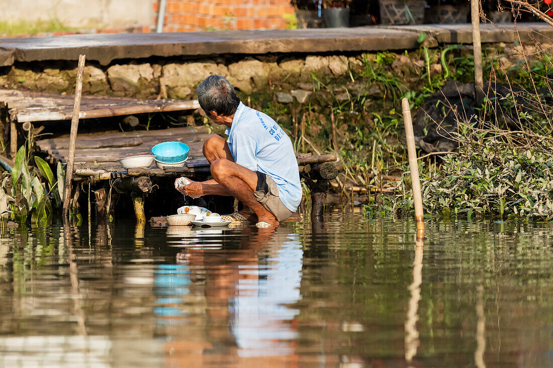 Mann reinigt Geschirr im Wasser des Hau-Flusses, schwimmender Markt von Cai Rang; Can Tho, Vietnam