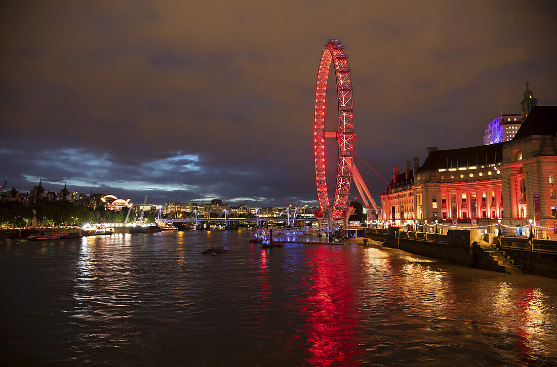 London Eye ferris wheel illuminated by red light on the shores of the River Thames; London, England
