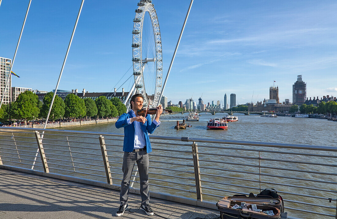Geige spielender Straßenmusiker auf einer Brücke über die Themse mit Booten und dem London Eye im Hintergrund; London, England