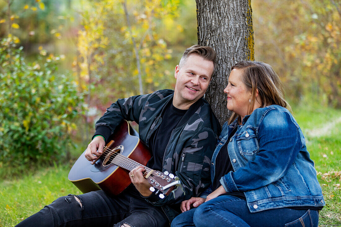 Ein reifes Paar, das viel Zeit miteinander verbringt. Die Frau hört ihrem Mann zu, wie er singt und Gitarre spielt, während sie an einem warmen Herbstabend in einem Stadtpark ist: Edmonton, Alberta, Kanada
