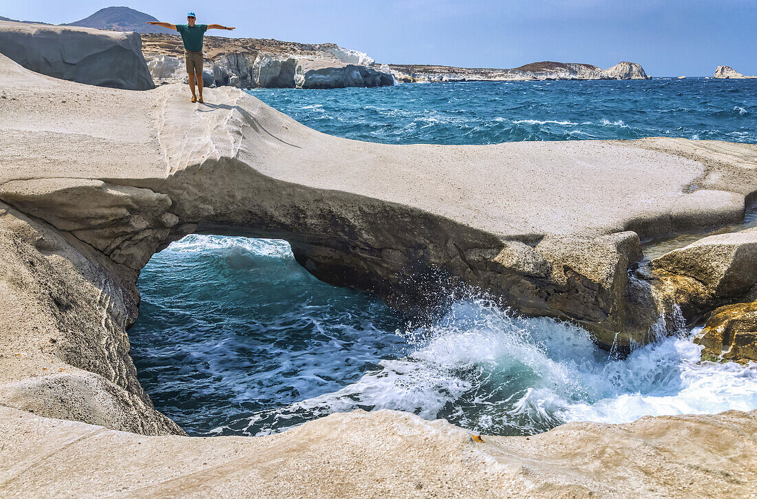 Männlicher Tourist steht auf einer einzigartigen Felsformation mit natürlichem Bogen und Brücke entlang der Küste von Milos, einer griechischen Insel; Milos, Griechenland