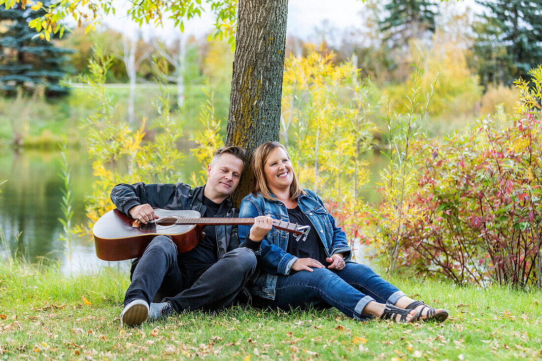 A mature couple spending quality time together and the wife is listening to her husband singing and playing his guitar while in a city park on a warm fall evening: Edmonton, Alberta, Canada