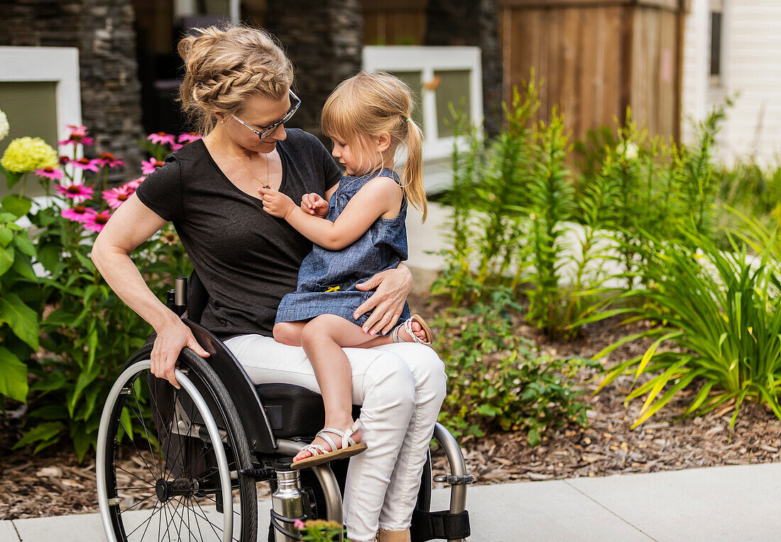 A paraplegic mom holding her little girl in her lap while sitting in her wheelchair in her front yard on a warm summer afternoon: Edmonton, Alberta, Canada.