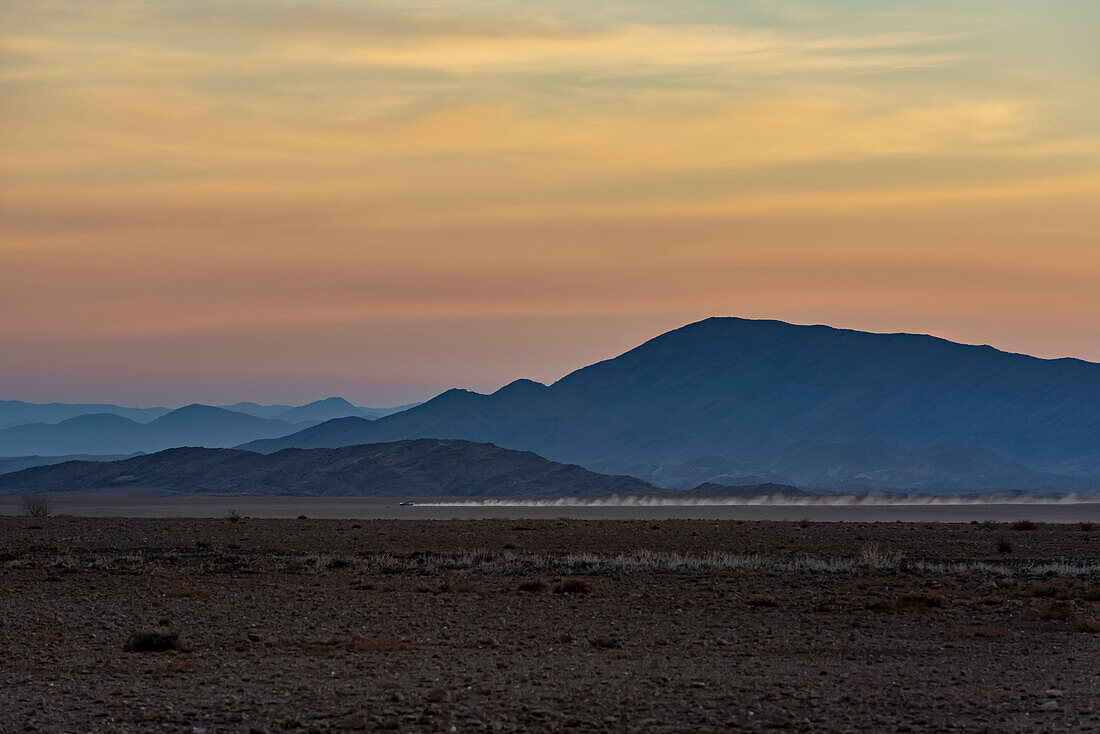 Sonnenaufgang in Aluvlei, Namib-Naukluft-Nationalpark, mit einem Auto, das über die Landschaft fährt und Staub in der Luft hinterlässt; Namibia