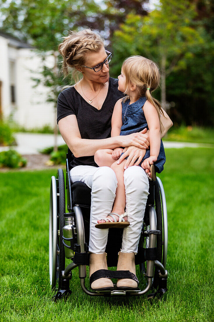 Eine querschnittsgelähmte Mutter hält ihr kleines Mädchen auf dem Schoß, während sie an einem warmen Sommernachmittag in ihrem Rollstuhl im Vorgarten sitzt: Edmonton, Alberta, Kanada.