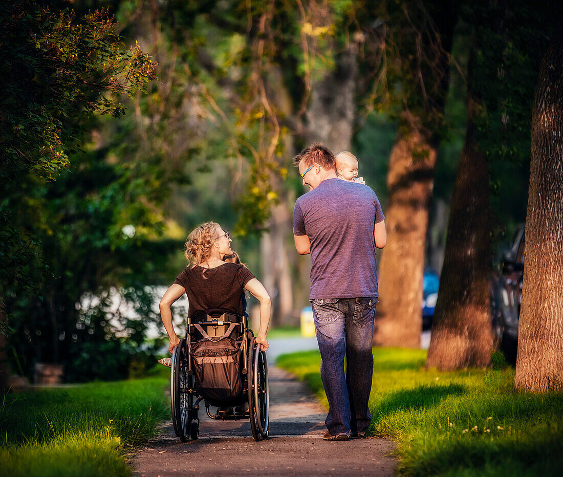 Blick von hinten auf eine Familie, die abends spazieren geht, und die Mutter ist querschnittsgelähmt und sitzt im Rollstuhl; Edmonton, Alberta, Kanada