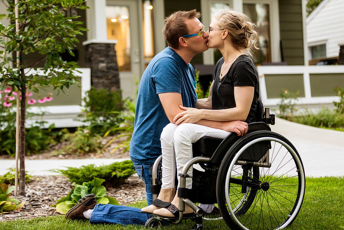 Ein Ehemann und seine querschnittsgelähmte Frau küssen sich an einem warmen Herbsttag in ihrem Vorgarten: Edmonton, Alberta, Kanada