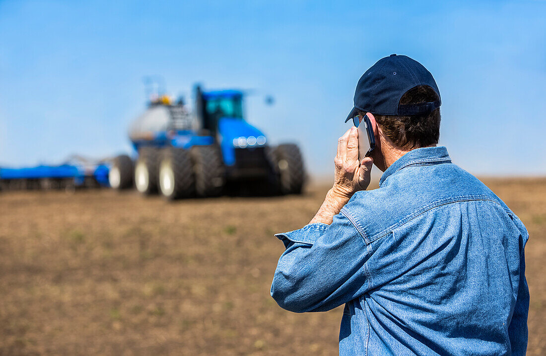 Landwirt benutzt ein Smartphone, während er auf einem landwirtschaftlichen Feld steht und den Traktor und die Geräte beim Säen des Feldes beobachtet; Alberta, Kanada