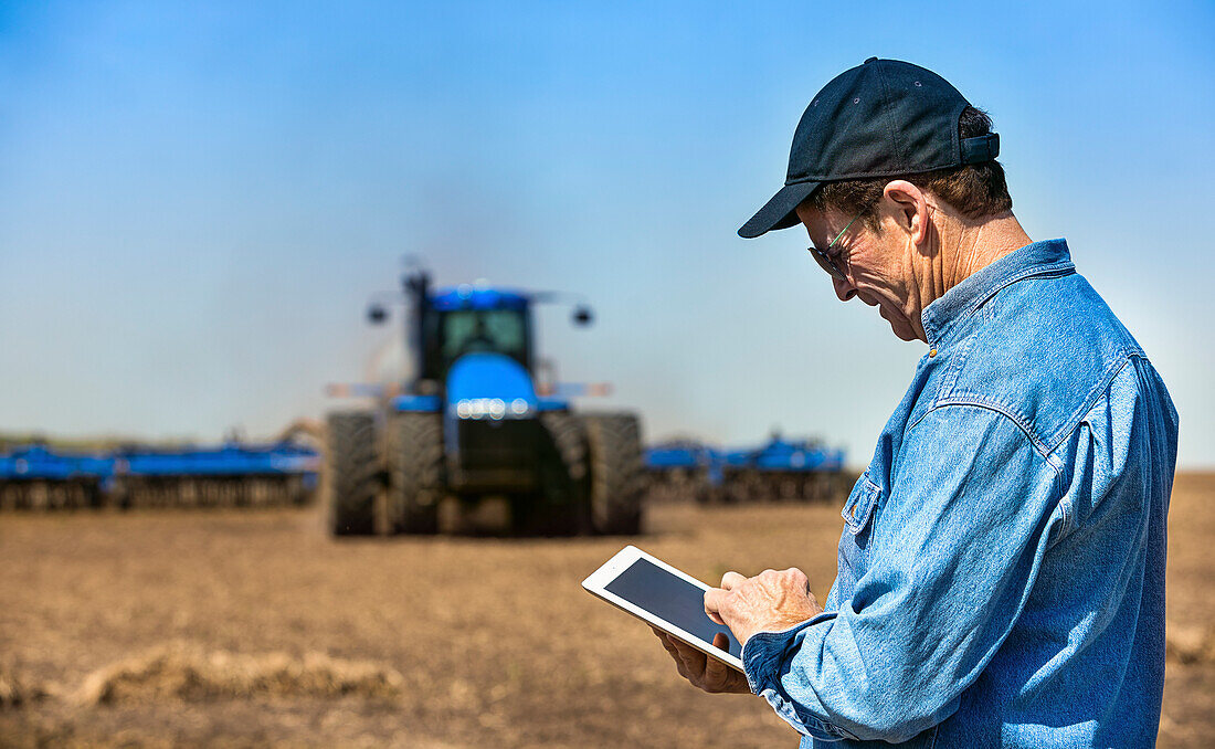 Farmer using a tablet while standing on a farm field and a tractor and equipment seeds the field; Alberta, Canada