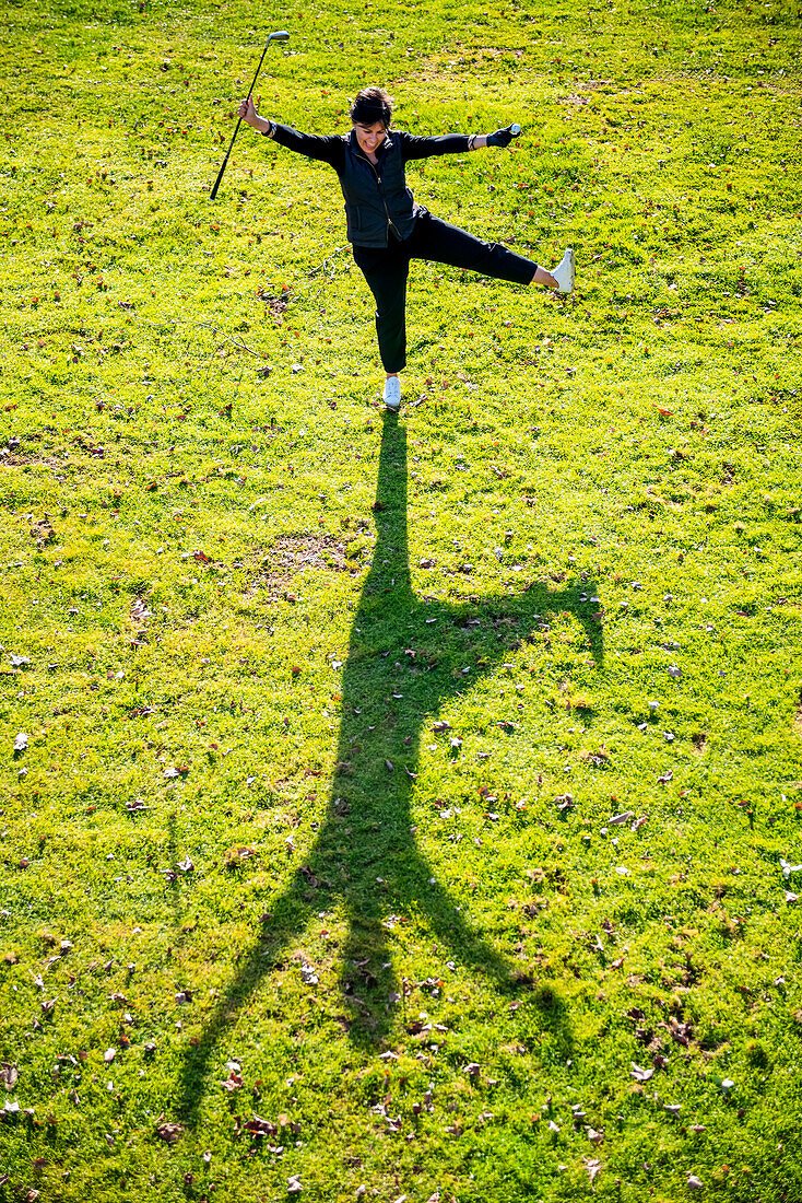 High angle view of female golfer on green grass holding her club and ball in the air and casting a shadow on the ground; Switzerland
