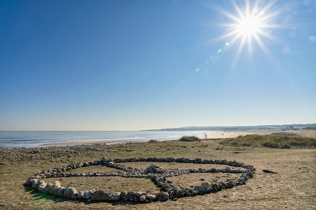 Symbolic rock formation of an X inside a circle made on the grass along the coast with people on the beach in the background; South Shields, Tyne and Wear, England