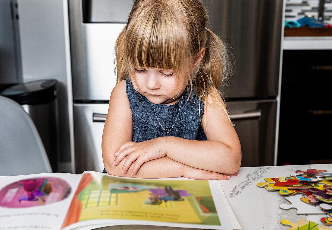 Ein süßes kleines Mädchen, das ein Bilderbuch liest und ein Puzzle auf einem Tisch in der Küche macht: Edmonton, Alberta, Kanada