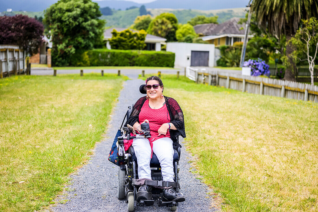 Maori-Frau mit Cerebralparese in einem Rollstuhl, die einen Bürgersteig hinunterfährt; Wellington, Neuseeland