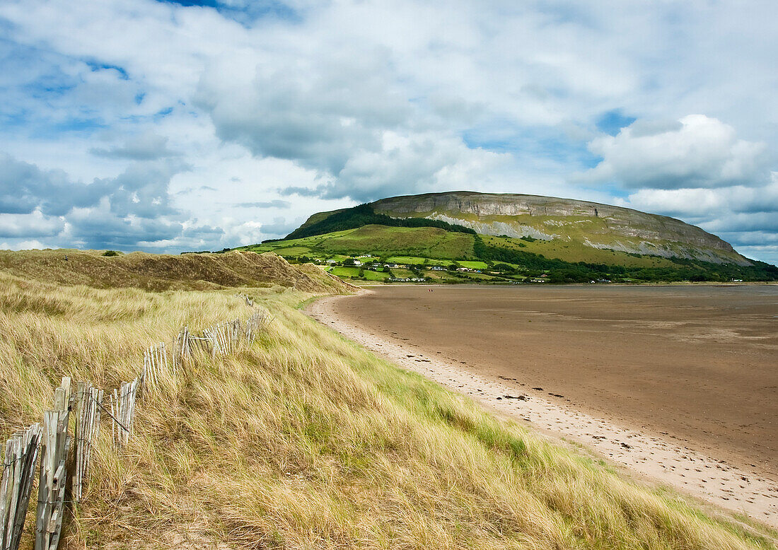 Irische Küste mit Strandgras, Ebbe und altem Holzzaun, mit einem Plateauberg und Klippen im Hintergrund im Sommer; Strandhill, County Sligo, Irland
