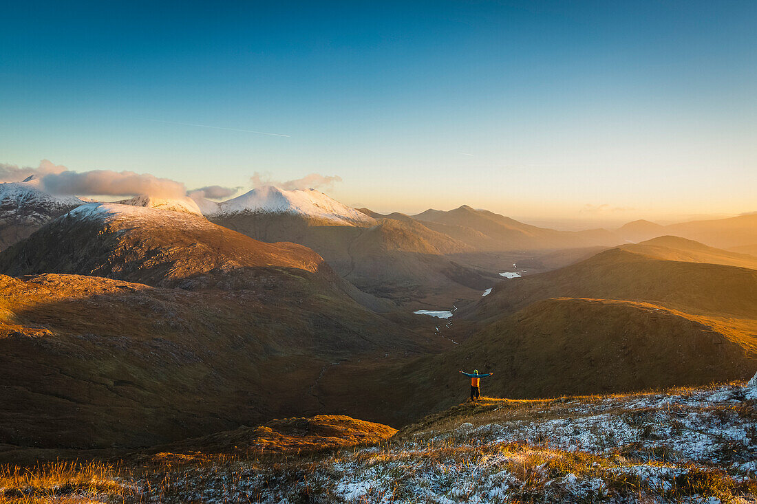 Mann mit ausgestreckten Armen an der Seite eines Berges mit Blick auf ein Tal und schneebedeckte Berge im Killarney National Park bei Sonnenaufgang; County Kerry, Irland