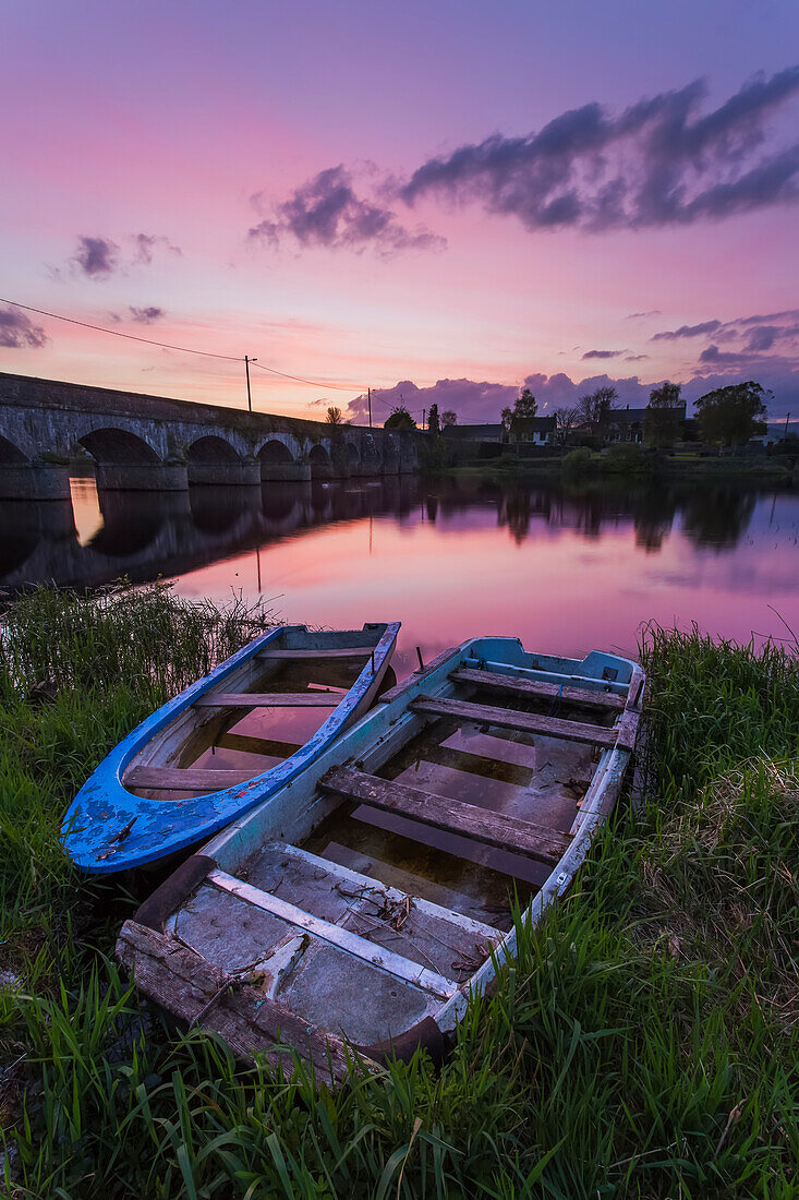 Zwei alte Holzboote am Ufer des Shannon River bei Sonnenuntergang mit einer Steinbrücke im Hintergrund; Montpellier, Grafschaft Limerick, Irland