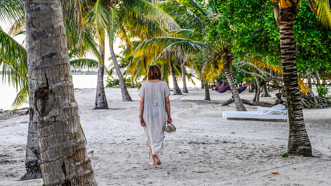 Eine reife Frau spaziert auf dem weißen Sand zwischen den Palmen an einem Strand in der Karibik, Placencia Peninsula; Belize