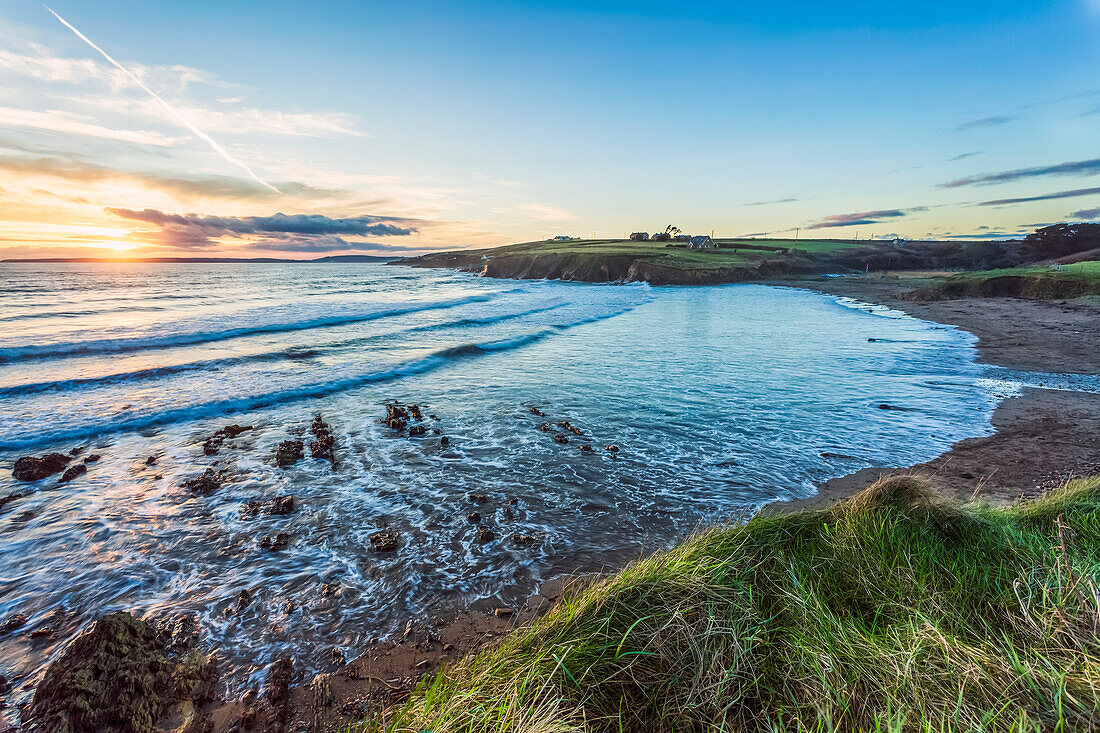 Sommerlicher Sonnenuntergang über der Bucht von Inch Beach mit Wellen im Meer und Gras im Vordergrund; County Cork, Irland