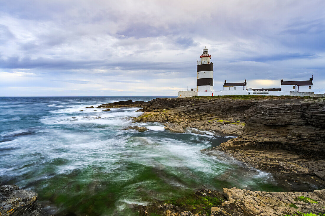 Hook Head lighthouse, Black and white lighthouse on rocky shore on a cloudy summer day., long exposure; County Waterford, Ireland