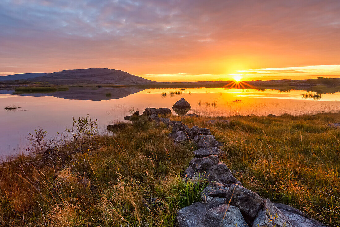 Small stone wall leading to a lake with a mountain and sunrise reflected in the water, Burren National Park; County Clare, Ireland