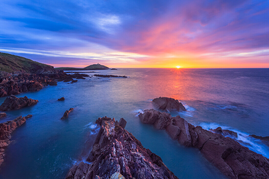 Sonnenaufgang über der irischen Küste mit zerklüfteten Felsen im Vordergrund und einem Leuchtturm auf einer Insel in der Ferne; Ballycotton, County Cork, Irland