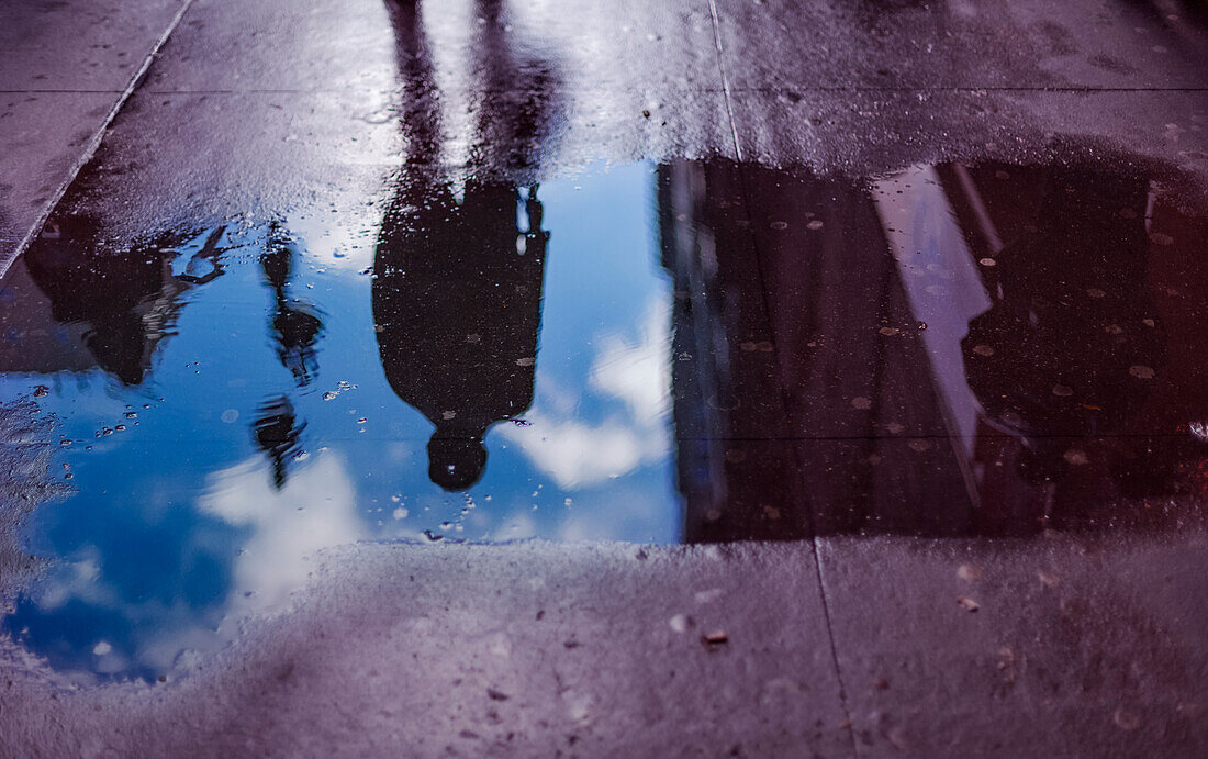 Die Figur eines Fußgängers und ein Wohnhaus spiegeln sich in einer Pfütze auf einem nassen Gehweg, Manhattan; New York City, New York, Vereinigte Staaten von Amerika