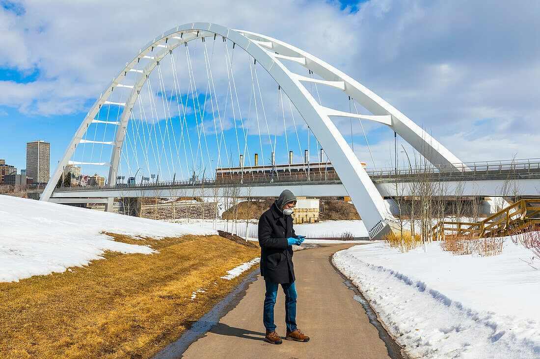 Ein Mann mit Maske und Handschuhen steht auf einem Weg im Freien und benutzt sein Smartphone während der Weltpandemie COVID-19; Edmonton, Alberta, Kanada