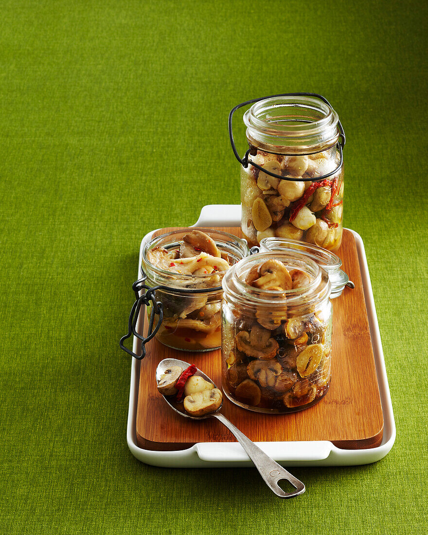 Preserved Mushrooms in Jars with Spoon on Wooden Cutting Board and Tray on Green Background in Studio