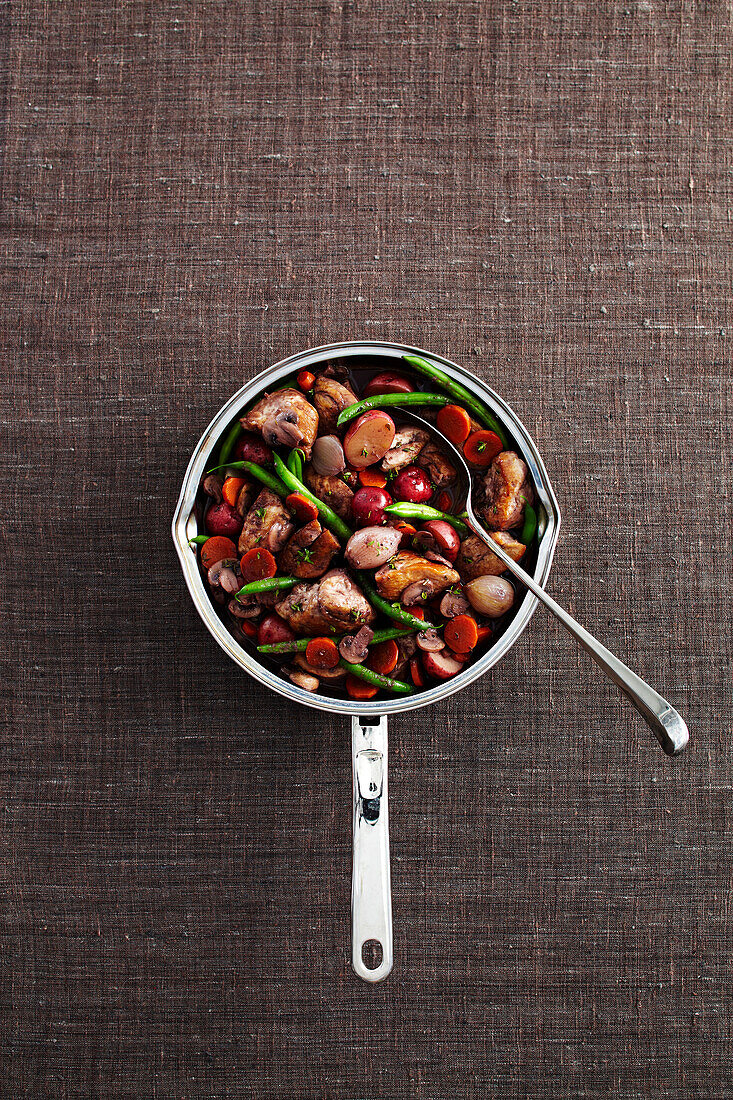Coq au vin in cooking pot with serving spoon on grey background, studio shot