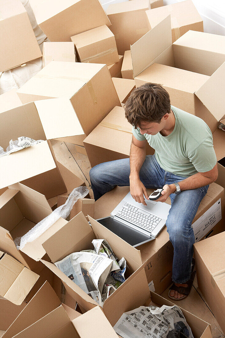 Man Sitting amongst Moving Boxes with Laptop Computer and Cellular Phone