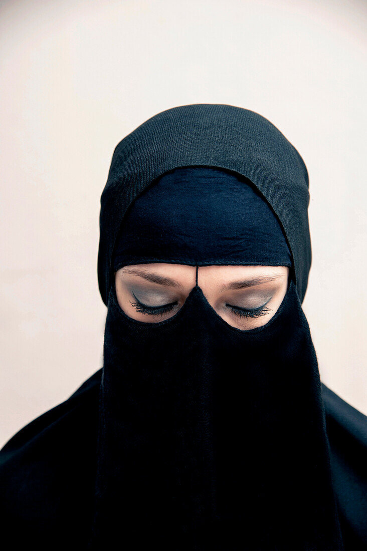 Close-up portrait of young woman wearing black, muslim hijab and muslim dress, eyes closed showing eye makeup, studio shot on white background