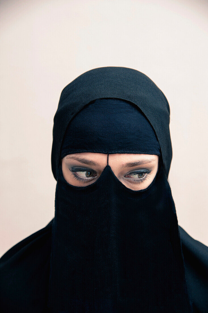 Close-up-Porträt einer jungen Frau, die einen schwarzen muslimischen Hidschab und ein muslimisches Kleid trägt, die Augen schauen zur Seite und zeigen Augen-Make-up, Studioaufnahme auf weißem Hintergrund